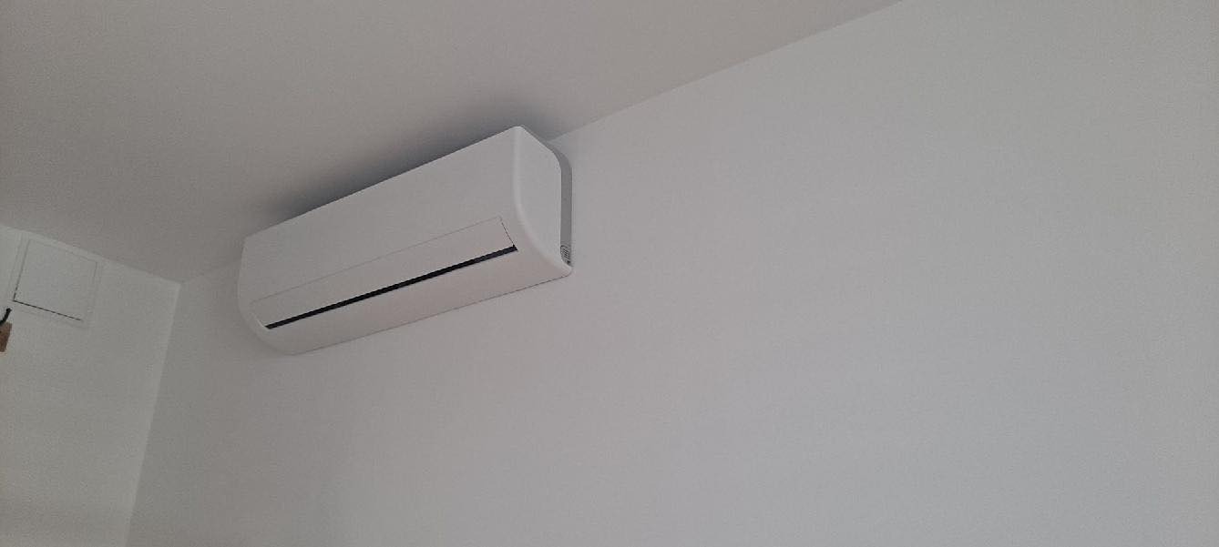 Biała jednostka wewnętrzna klimatyzacji zamontowana na białej ścianie w rogu pomieszczenia, widoczny fragment skrzynki elektrycznej po lewej stronie.
