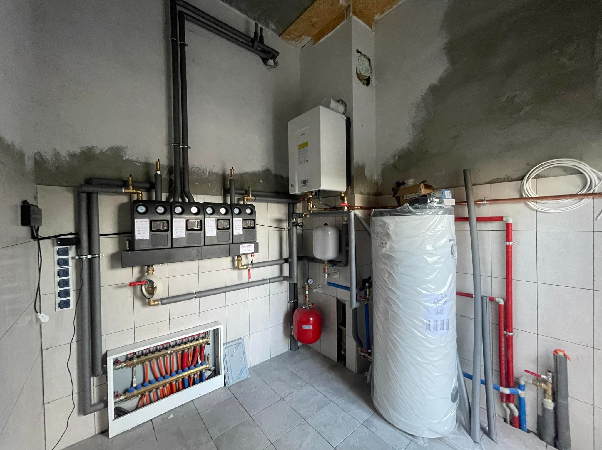 Kompletna instalacja grzewcza w kotłowni: rozdzielacz ogrzewania podłogowego z licznymi pętlami, zbiornik buforowy owinięty folią, ekspansomat, piec gazowy na ścianie, rury i zawory na tle surowych...