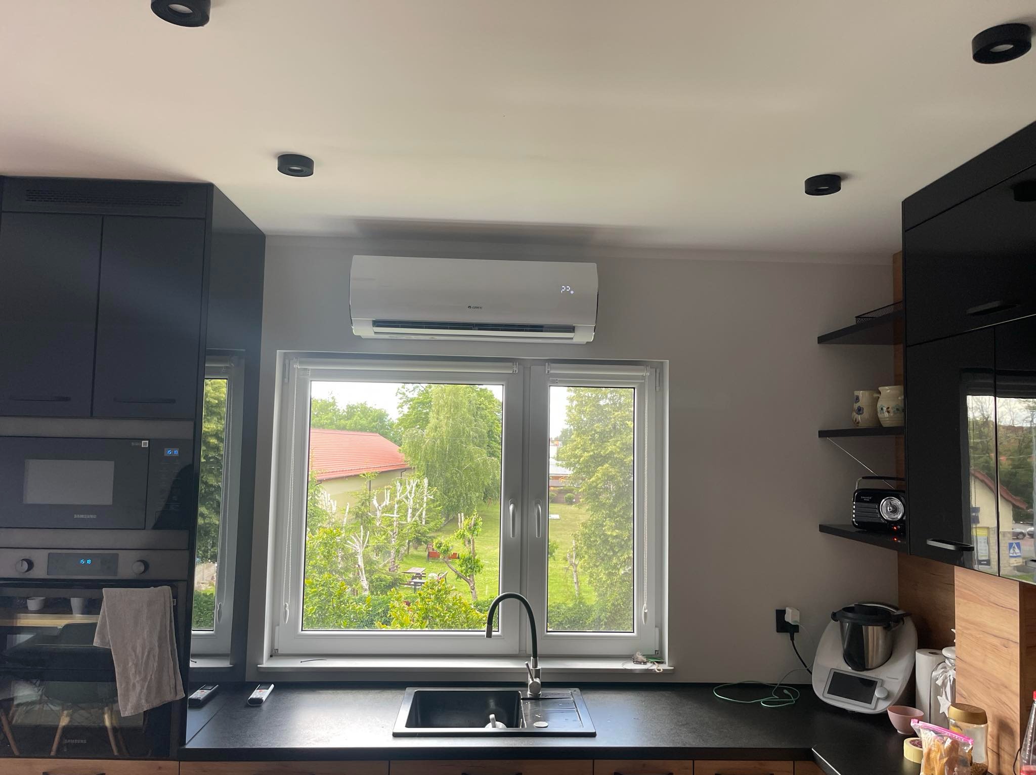 Klimatyzator Gree zamontowany nad oknem w kuchni z widokiem na ogród, wyświetlający temperaturę 22 stopnie, z czarnymi szafkami kuchennymi po bokach.