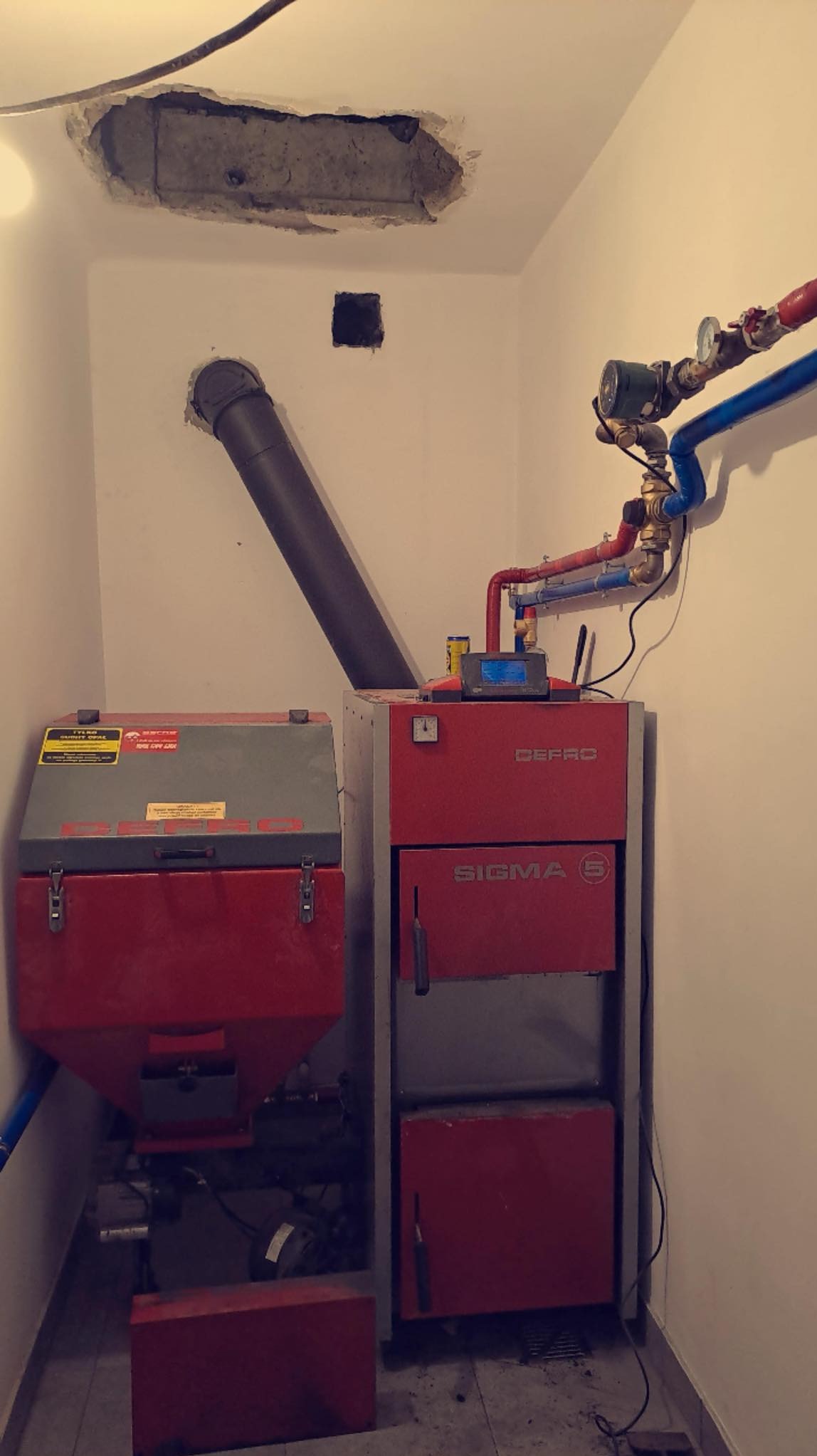 Kotłownia z dwoma piecami na paliwo stałe marki DEFRO SIGMA z widocznymi instalacjami hydraulicznymi, rurą odprowadzającą spaliny i niedokończonym sufitem w pomieszczeniu.
