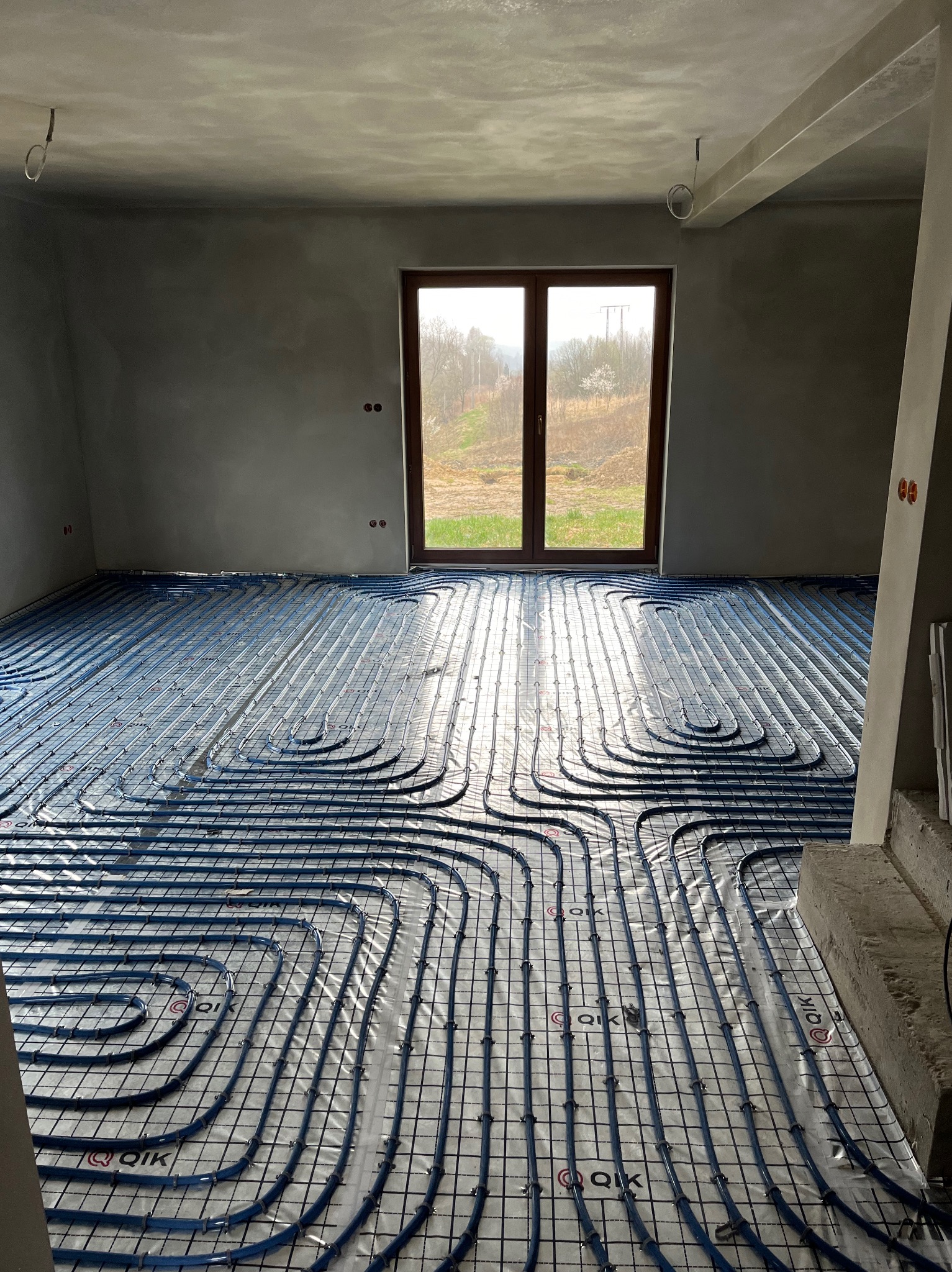 Niedokończona instalacja ogrzewania podłogowego z niebieskimi rurami na folii w surowym, jasnym pomieszczeniu z dużym oknem i widokiem na drzewa.