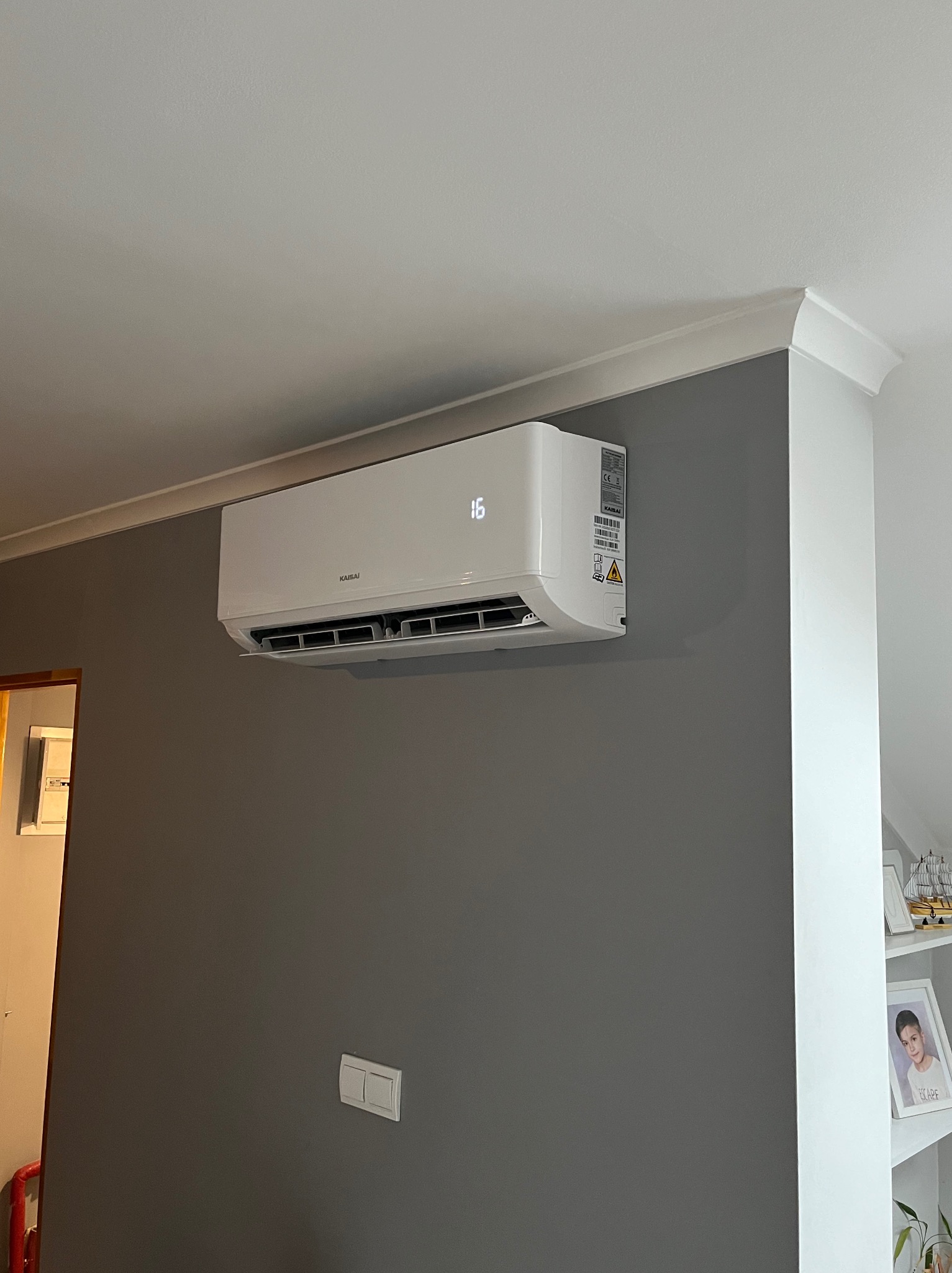 Biała jednostka klimatyzacji Kaisai zamontowana na szarej ścianie w pomieszczeniu, widoczny wyświetlacz z temperaturą 16 stopni, listwa sufitowa i włącznik światła.