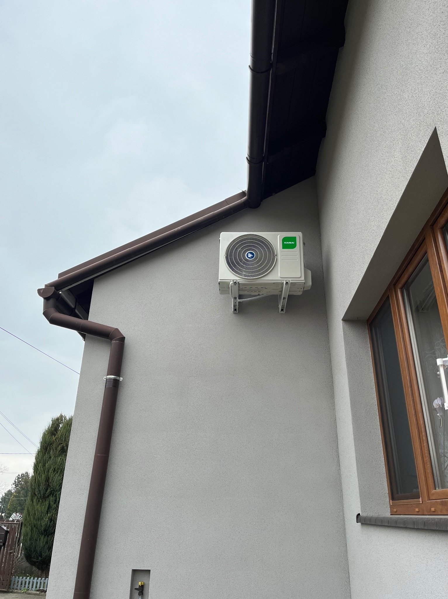 Zewnętrzna jednostka klimatyzacji Kaisai zamontowana na szarej elewacji domu, widoczny brązowy system rynnowy i okno z drewnianą ramą w pochmurny dzień.