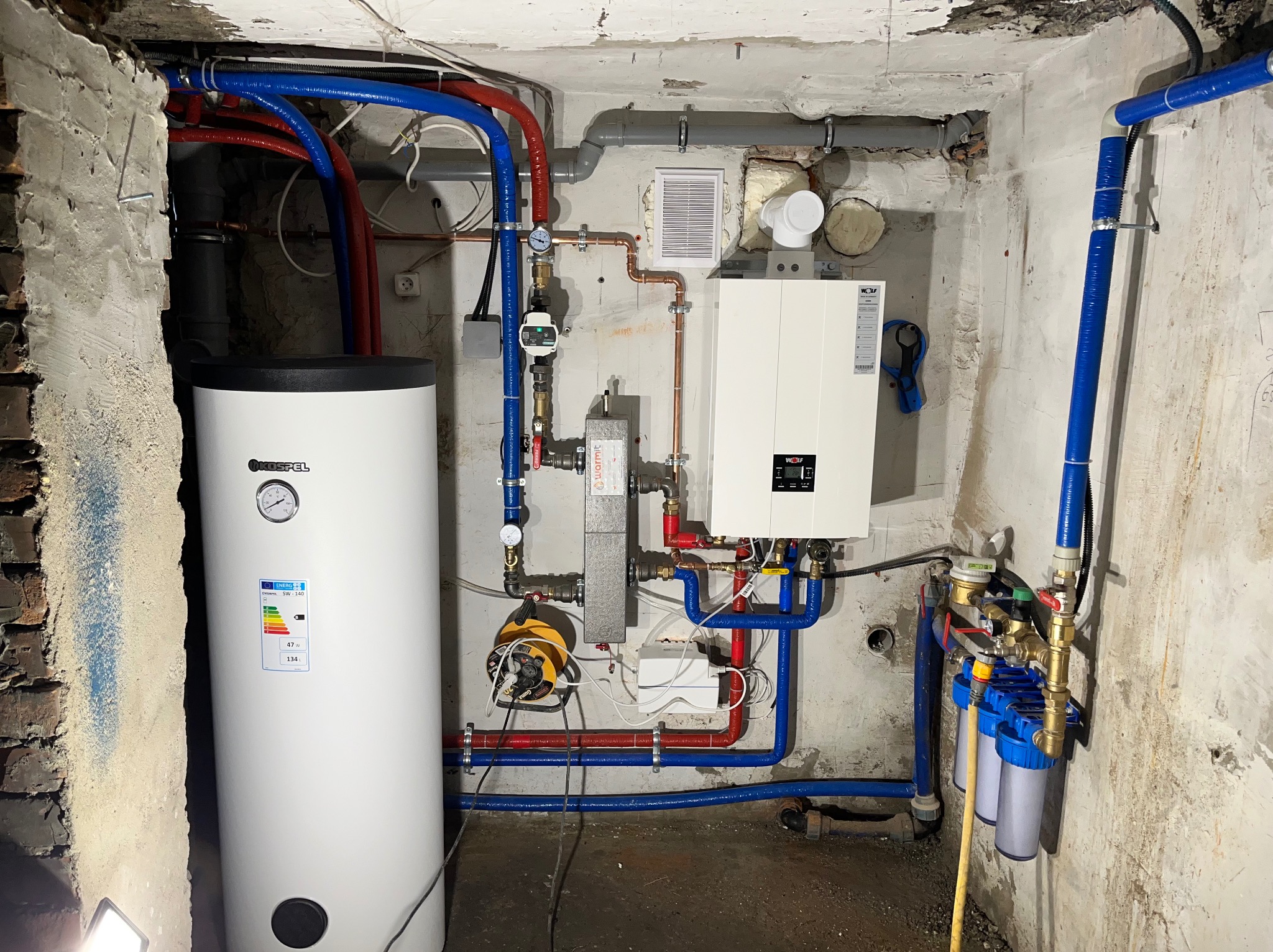 Zainstalowany biały piec gazowy marki Wolf z podłączonymi rurami miedzianymi i niebieskimi rurami z tworzywa oraz biały zasobnik ciepłej wody marki Kospel w surowym pomieszczeniu kotłowni.