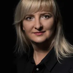 Portret kobiety z blond włosami, ubrana w czarną koszulę, na ciemnym tle.