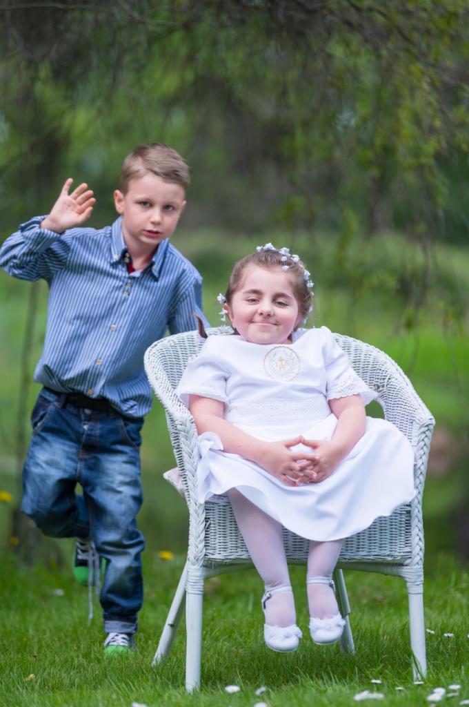 Dziewczynka w białej sukience komunijnej siedzi na wiklinowym krześle, obok stoi chłopiec w niebieskiej koszuli na tle zieleni ogrodu.