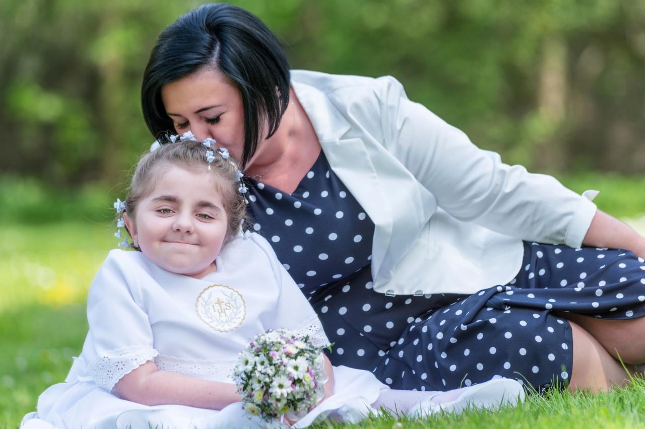 Mama całuje w głowę uśmiechniętą dziewczynkę w białej sukience komunijnej z bukiecikiem na trawie.