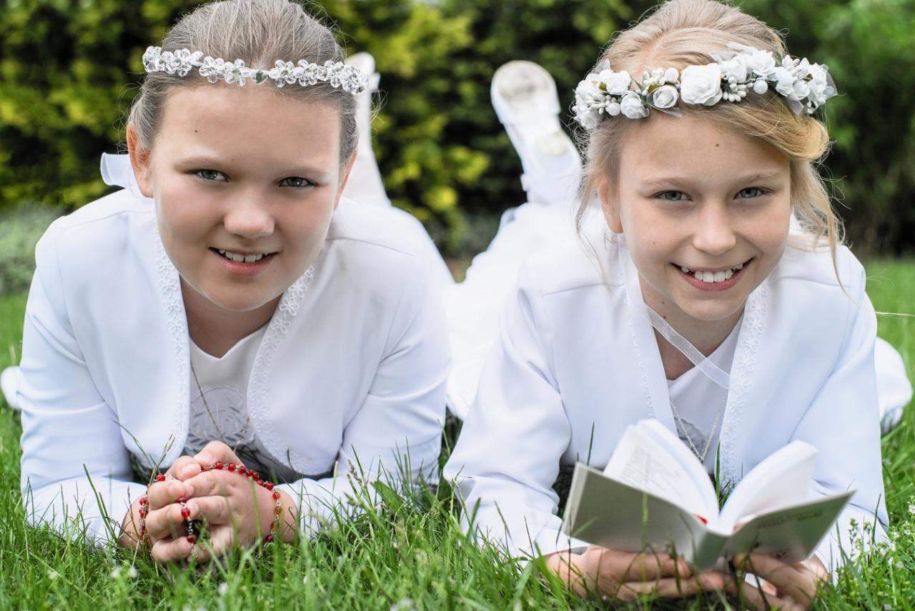 Dwie dziewczynki w białych strojach komunijnych leżą na trawie, jedna trzyma różaniec, druga czyta książkę, obie uśmiechnięte.