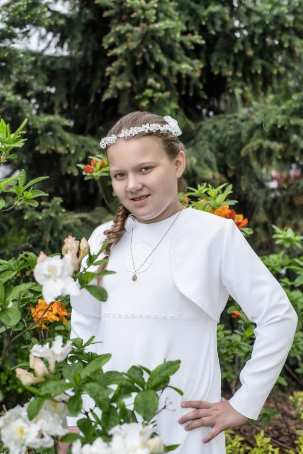 Dziewczynka w białej sukience komunijnej, z wiankiem na głowie i medalikiem na szyi, pozuje na tle zieleni i kwiatów.