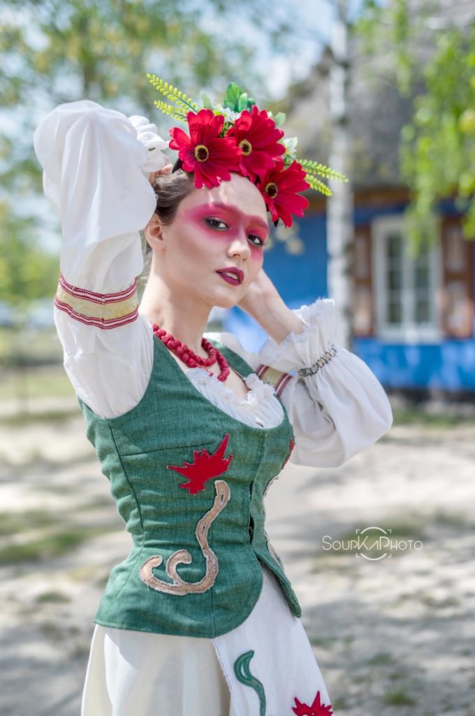 Kobieta w ludowym stroju z wiankiem z czerwonych kwiatów na głowie, pozująca na tle drewnianego domu w Łodzi. Nietypowy makijaż z czerwonymi cieniami na powiekach.