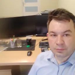 Mężczyzna w jasnoniebieskiej koszuli siedzi za biurkiem z dwoma monitorami, kalkulatorem, myszką, kubkiem i szklanką wody.