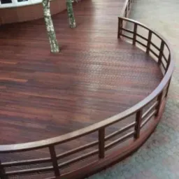 Zewnętrzny taras z ciemnego drewna z zaokrągloną balustradą i wkomponowanymi drzewami, widok z góry na fragment chodnika i osoby w oddali.