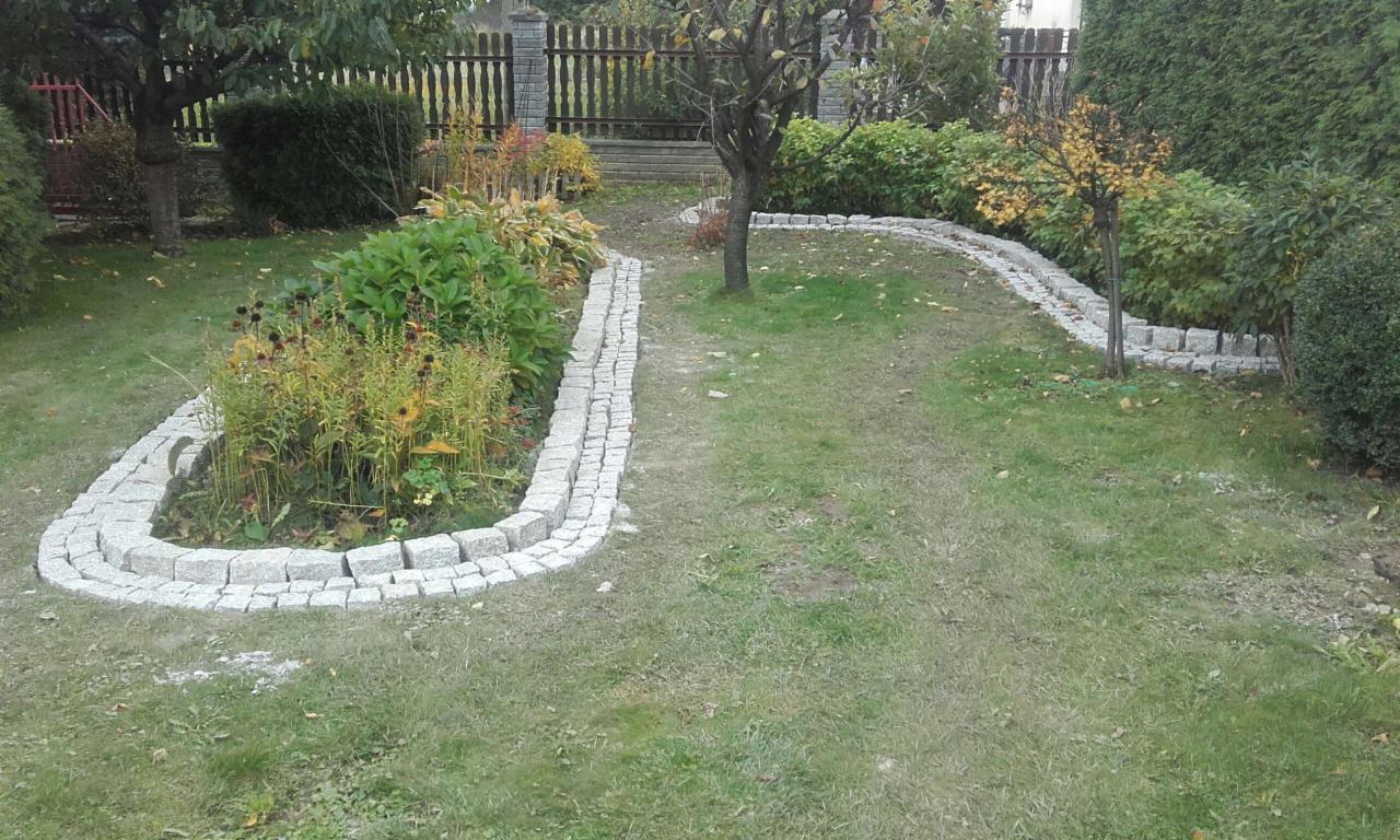 Krawężniki z kostki granitowej wyznaczające ścieżkę i obrzeża rabat w ogrodzie z drzewami i krzewami w tle.