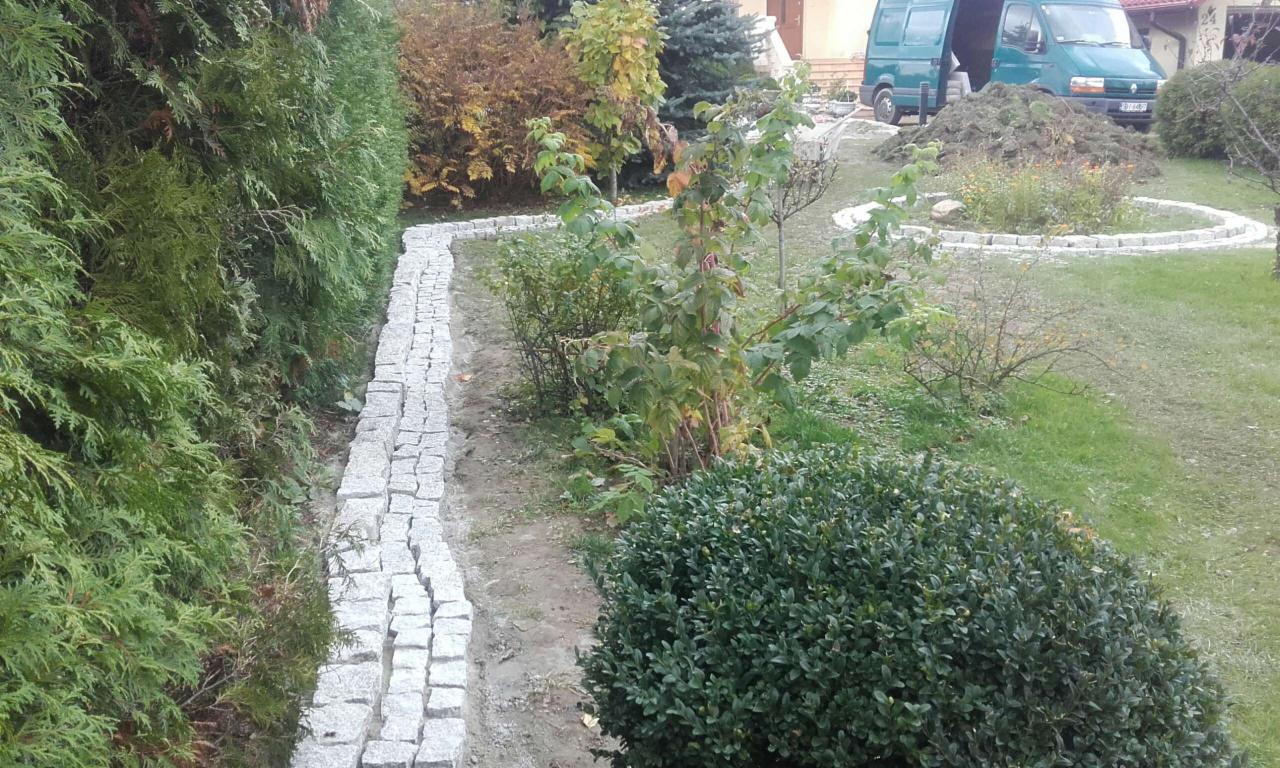 Krawężnik z kostki granitowej oddzielający rabatę z tujami od trawnika, w tle zaparkowany zielony bus i świeżo nasadzona roślinność w okręgu z obrzeży.