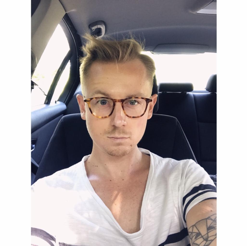 Mężczyzna z blond włosami i okularami w brązowych oprawkach, siedzący w samochodzie, z tatuażem geometrycznym na ramieniu i koszulce w paski.