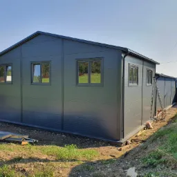 Gotowy budynek modułowy z płyt warstwowych w kolorze grafitowym, widok z zewnątrz, z widocznymi oknami i fragmentami terenu przed budynkiem.