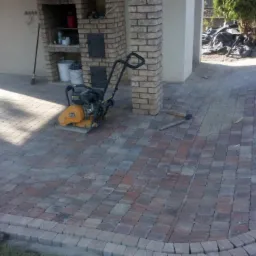 Układanie kostki brukowej na tarasie, zagęszczarka spalinowa obok kolumny z cegły, widoczne narzędzia i przygotowane podłoże.