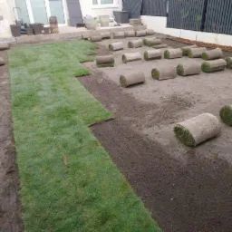 Fragment układania trawy w rolkach na przygotowanym podłożu, widoczne rozłożone pasy i zwinięte rolki, narzędzie ogrodnicze w prawym dolnym rogu.