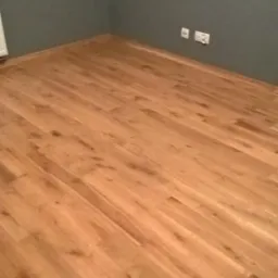 Wykończony pokój z panelami podłogowymi w odcieniach brązu, szare ściany z gniazdkami elektrycznymi, widok na narożnik pomieszczenia.