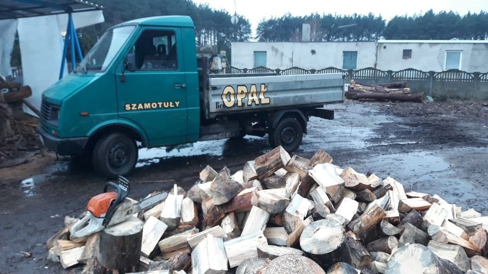 Dostawa drewna opałowego w Piotrowie: zielony samochód dostawczy z napisem 'OPAŁ' zaparkowany obok stosu pociętego drewna, na pniu leży piła spalinowa.