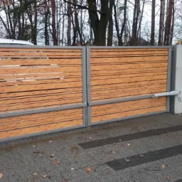 Drewniana brama przesuwna z automatycznym napędem, widoczne siłowniki, szara metalowa rama i betonowe słupki, na podjeździe z kostki brukowej.