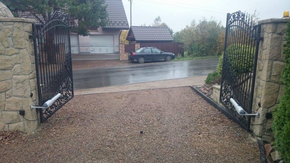 Otwarta, dwuskrzydłowa brama z czarnego metalu z automatycznym napędem, zamontowana na kamiennych słupkach, widok na podjazd z żwiru i mokrą drogę, w tle dom z garażem i zaparkowany samochód.