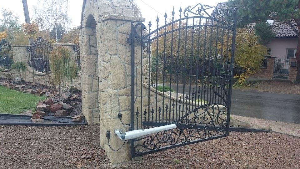 Otwarta, ozdobna, czarna brama wjazdowa z kamiennymi słupkami, wyposażona w biały siłownik, na tle posesji z ogrodem.