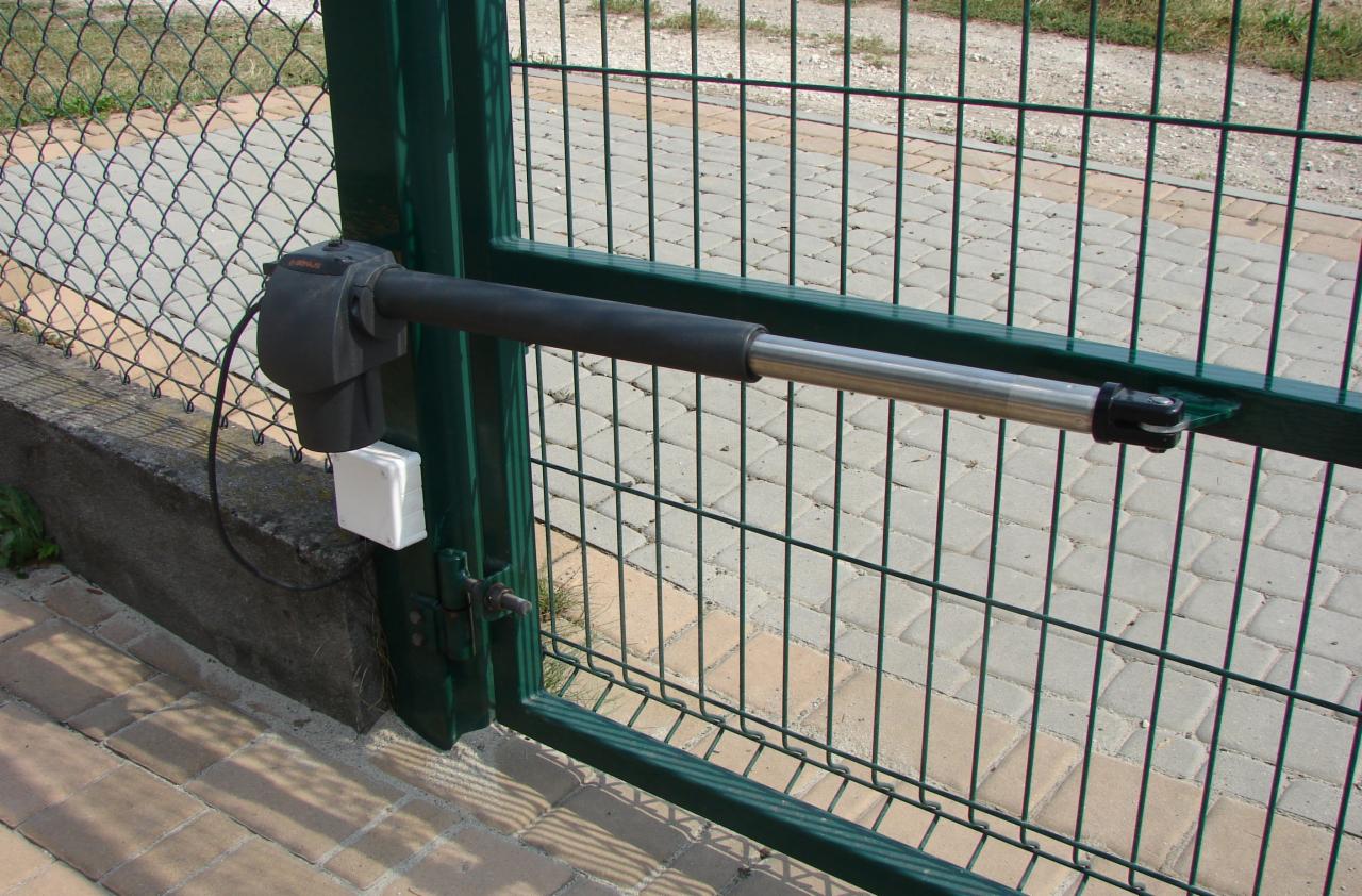 Zamknięta brama z siatki z zamontowanym siłownikiem ramieniowym, widoczna kostka brukowa i trawa w tle.