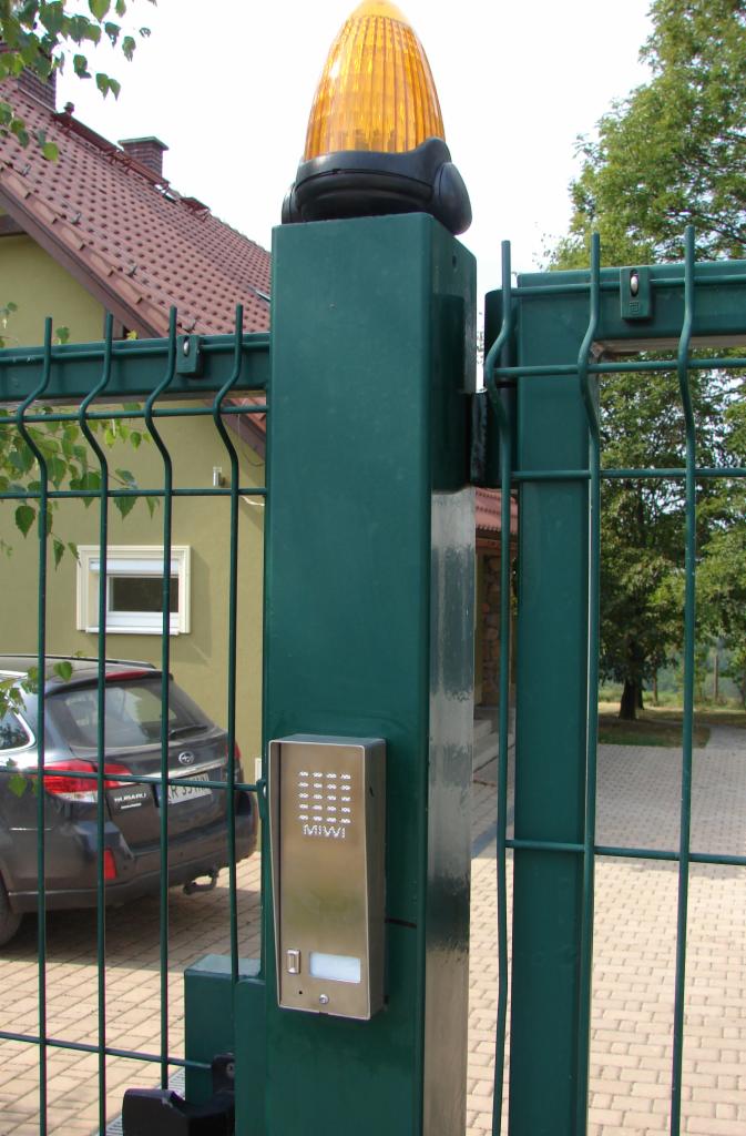 Zielona brama wjazdowa z panelem kodowym MIWI i pomarańczową lampą ostrzegawczą na słupku, widoczny dom i zaparkowany samochód.