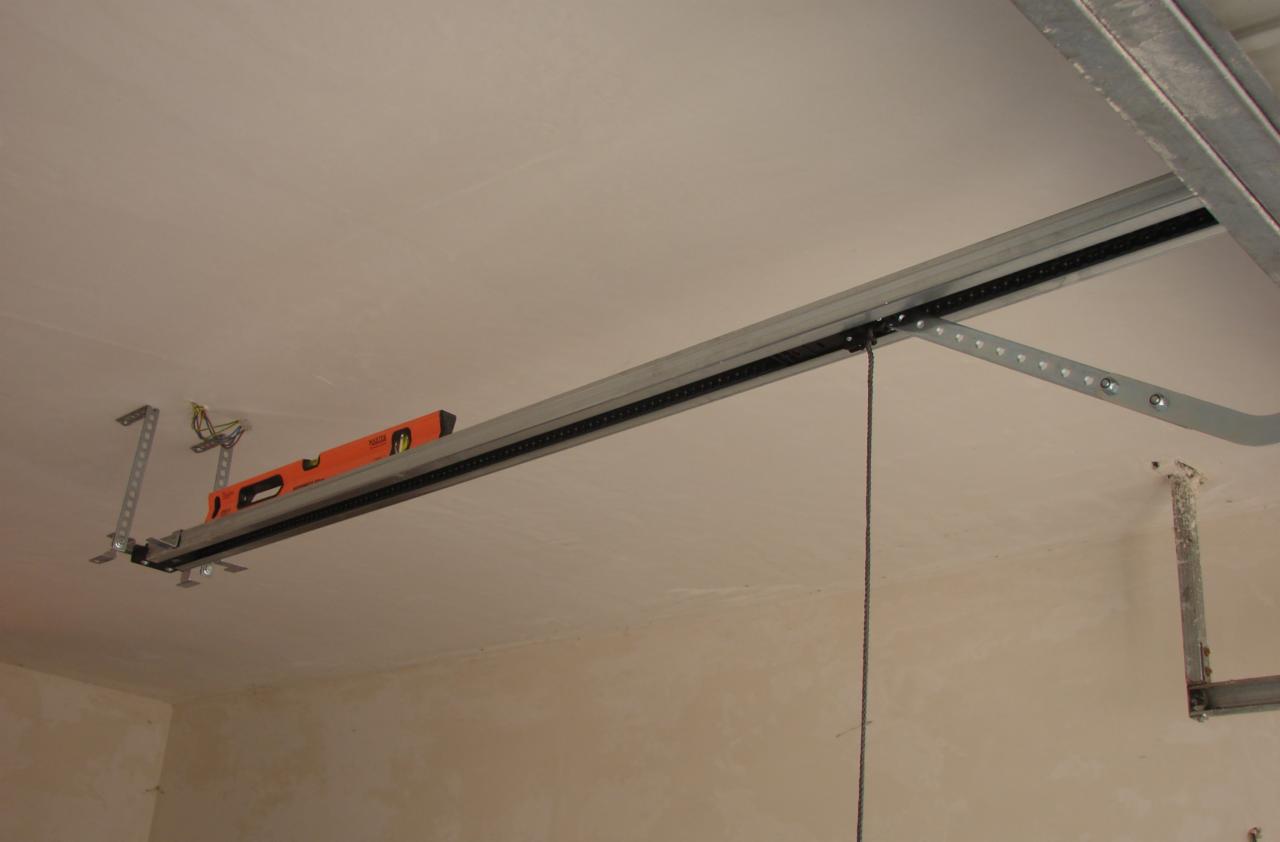 Montaż automatyki bramy garażowej: szyna z łańcuchem podwieszona do sufitu, poziomica na szynie, widoczne przewody elektryczne.