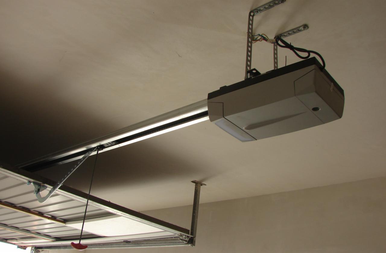 Zamontowany napęd bramy garażowej pod sufitem, widoczne mocowania i szyna prowadząca, fragment bramy garażowej z linką awaryjną.