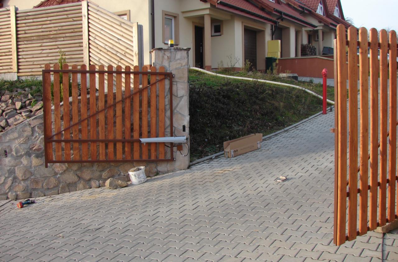 Drewniana brama skrzydłowa z zamontowanym napędem, widoczna skrzynka po napędzie, kostka brukowa i narzędzia podczas montażu.
