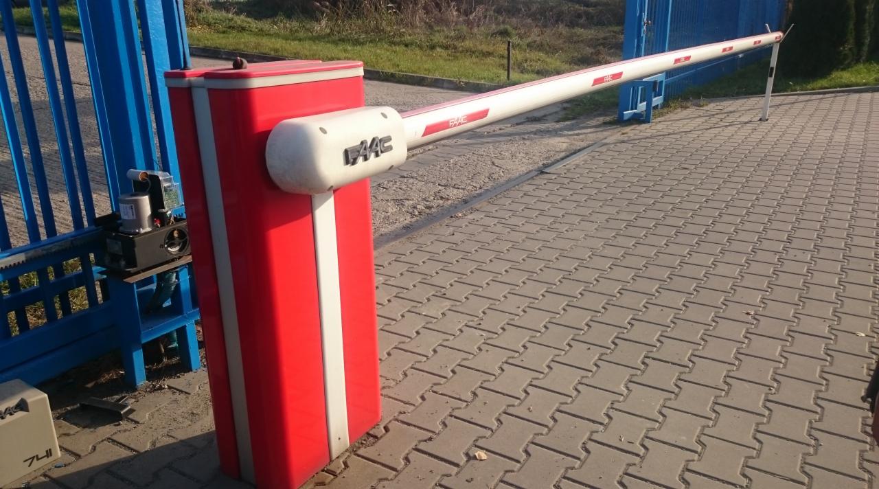 Szlaban parkingowy FAAC z czerwoną obudową i biało-czerwonym ramieniem, ustawiony na kostce brukowej, widoczny mechanizm sterujący przy niebieskiej bramie.