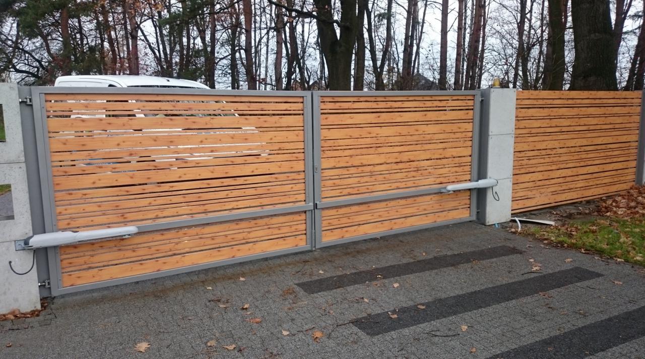 Drewniana brama przesuwna z automatycznym napędem, widoczne siłowniki, szara metalowa rama i betonowe słupki, na podjeździe z kostki brukowej.