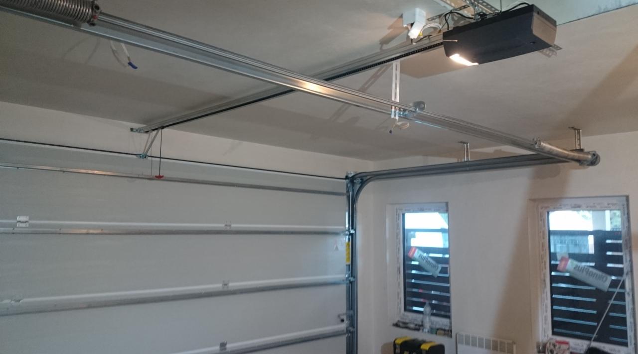 Widok od wewnątrz na zamontowaną automatykę do bram garażowych z widocznym napędem, szynami prowadzącymi i sprężyną, biała brama segmentowa w pozycji otwartej, dwa okna z roletami w tle.