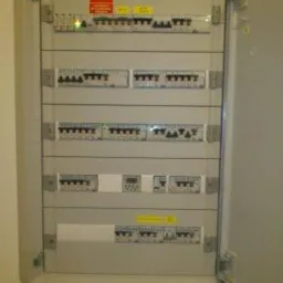 Otwarta skrzynka rozdzielcza z bezpiecznikami, zabezpieczeniami i licznikiem energii wewnątrz, widoczna naklejka ostrzegawcza.