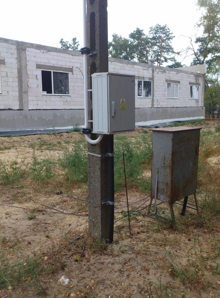 Skrzynka elektryczna z symbolem ostrzegawczym na słupie betonowym, obok metalowa skrzynka na stojaku, w tle budynek w stanie surowym.