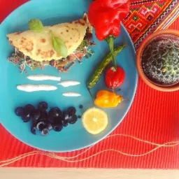 Turkusowy talerz z tortillą wypełnioną mięsem i warzywami, udekorowaną bazylią, obok oliwki, plasterek cytryny, krople sosu, papryczki chili i czerwona papryka, w tle kaktus i etniczny wzór.