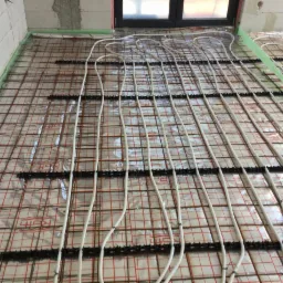 Montaż ogrzewania podłogowego: białe rurki grzewcze ułożone na metalowej siatce z folią izolacyjną, widoczny fragment ściany z bloczków.