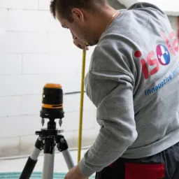 Pracownik w szarej bluzie z logo firmy ISOLA mierzy wysokość ż&oacute;łtą miarką, obok stoi poziomica laserowa na statywie, w tle ściana z białych bloczk&oacute;w.