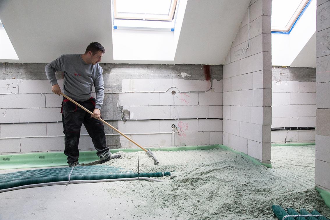 Pracownik rozprowadza jastrych na ogrzewaniu podłogowym w pomieszczeniu z surowymi ścianami z bloczków i oknami dachowymi.