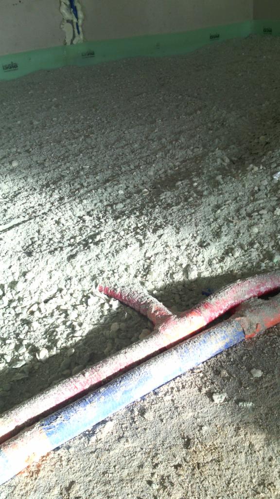Niedokończona podłoga z widocznymi rurami ogrzewania podłogowego, pokryta warstwą jastrychu przed wylaniem, zabezpieczona folią izolacyjną przy ścianach.