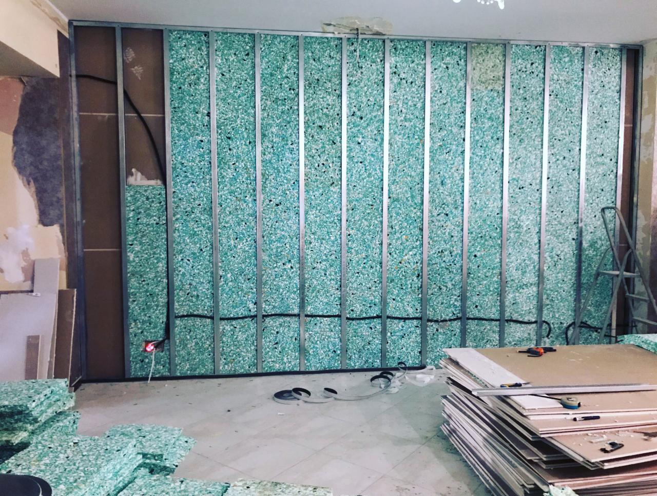 Montaż izolacji akustycznej z zielonej wełny mineralnej w metalowym stelażu na ścianie w trakcie remontu, widoczne kable elektryczne i narzędzia.