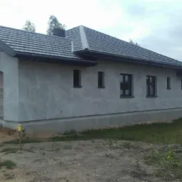 Dom jednorodzinny w trakcie tynkowania elewacji zewnętrznej, widoczne surowe wykończenie ścian i przygotowane podłoże.