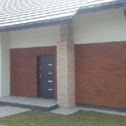 Nowoczesny dom jednorodzinny z elewacją w kolorze jasnego drewna, fragment ściany z oknem i ciemnymi drzwiami wejściowymi, wykończenie z kamienia dekoracyjnego na rogu budynku.