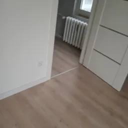 Jasne panele podłogowe ułożone w nowym pokoju, widoczny fragment ściany z gniazdkiem elektrycznym i drzwi z trzema poziomymi wgłębieniami, w tle grzejnik pod oknem.