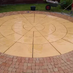 Okrągły plac z ułożoną żółtą kostką brukową, obramowany czerwoną kostką, widok z góry, częściowo mokra powierzchnia, w tle trawnik, taczka i narzędzia.