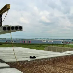 Montaż prefabrykowanych płyt stropowych z otworami, widoczni pracownicy w kamizelkach odblaskowych, zbrojenie stropu, krajobraz z halami przemysłowymi w tle.