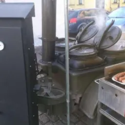 Polowa kuchnia z parującymi kotłami i talerzem grillowanych kiełbas na tle miejskiego krajobrazu.