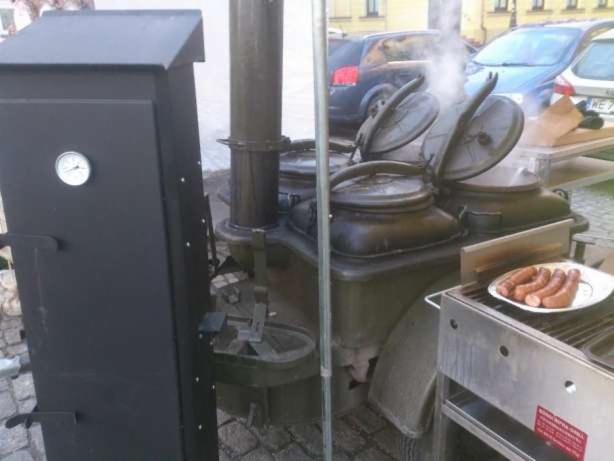 Polowa kuchnia z parującymi kotłami i talerzem grillowanych kiełbas na tle miejskiego krajobrazu.